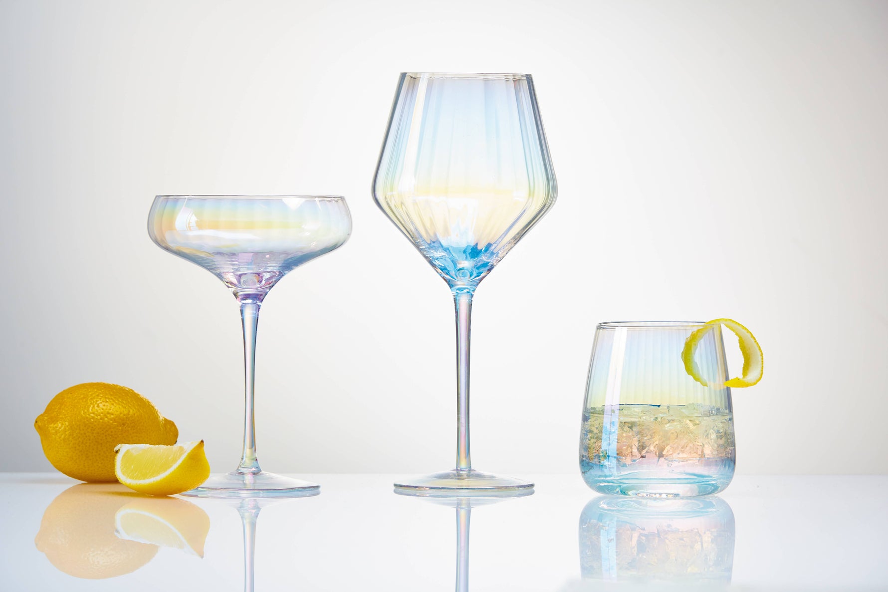 Product photography featuring three iridescent ribbed cocktail glasses including a coupe, a tall wine glass, and a tumblr with ice and a lemon twist, alongside a whole lemon and a wedge on a reflective white surface