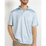A smiling man with a beard models a light blue and white short-sleeve polo shirt featuring a repeating stylized crab pattern, his hand casually in the pocket of khaki pants against a clean white studio background