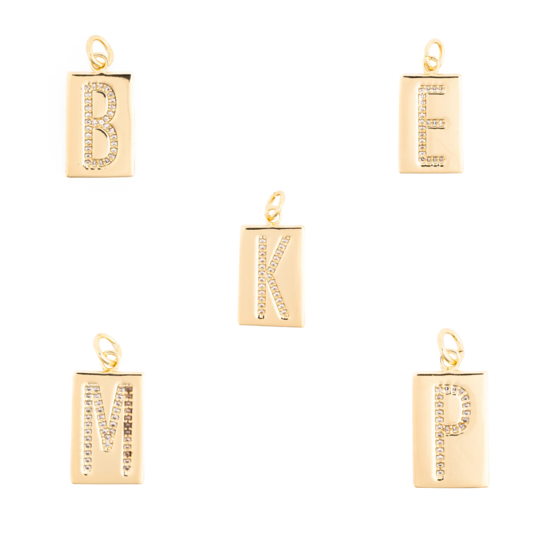 Jewelry design featuring five gold-toned rectangular alphabet pendants with pavé crystal initials B, E, K, M, and P on a white studio background with polished metallic finishes and top hanging bails