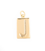 Gold jewelry pendant featuring a rectangular tag with a pave-set crystal letter J and a polished gold finish against a white studio background