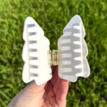 A hand holds a white bow-shaped hair claw clip with subtle pink accents and a clear gold-toned spring mechanism against a vibrant green grass background