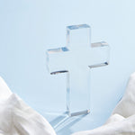 Clear crystal cross centered on a light blue background with a white textured cloth draped below, featuring sharp reflections and soft shadows