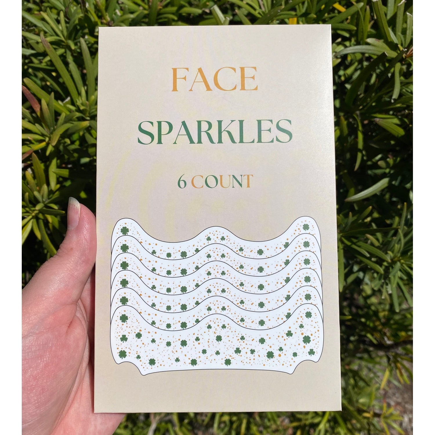 Cosmetic product package for Face Sparkles 6 Count, held in a hand outdoors, displays orange FACE and green SPARKLES text over a wavy white design sprinkled with green shamrocks and golden dots against a blurred green foliage background