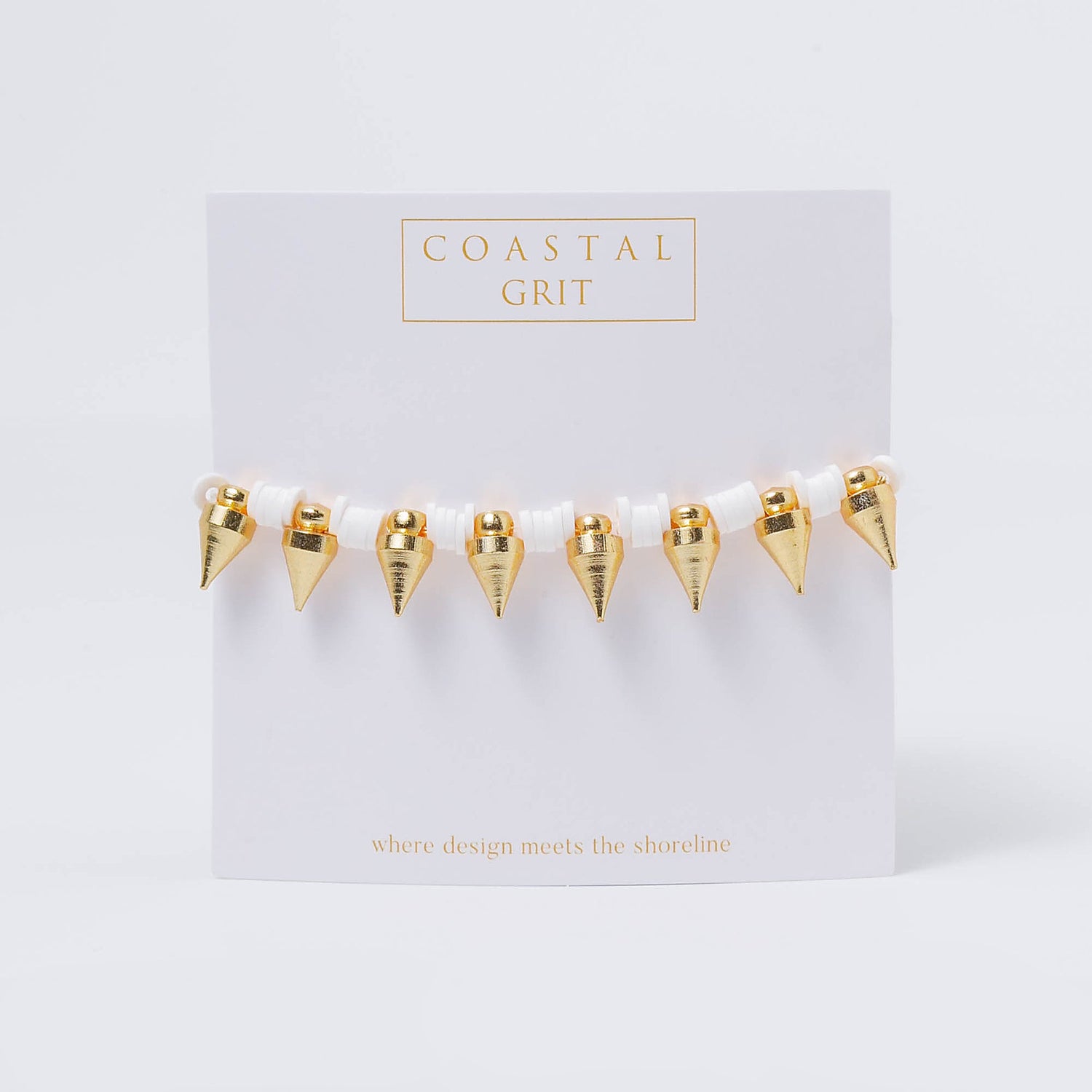 Product photography of a Coastal Grit bracelet on a white display card, featuring white disc beads and seven shiny gold cone-shaped charms, with the brand name Coastal Grit and its tagline where design meets the shoreline in gold lettering