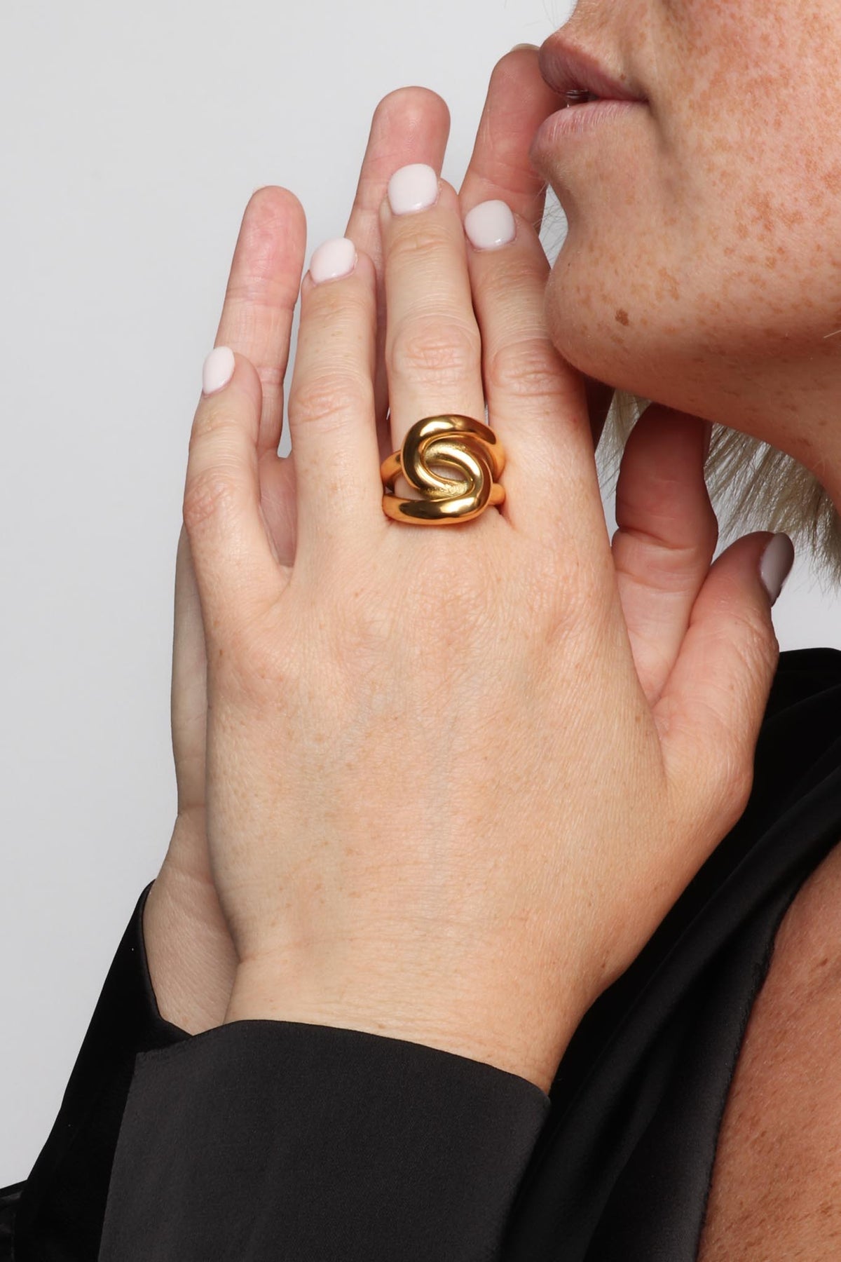 Woman's hand wearing a bold sculptural gold ring with pale pink nails held thoughtfully near her freckled mouth