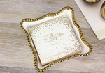 Photograph of white napkins with gold confetti and Cheers script stacked in a white scalloped tray with a beaded gold rim placed on a light wood grain table