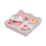 White ceramic napkin holder with a wavy edge filled with pink floral paper napkins tied with a white satin ribbon featuring vibrant pink and yellow dahlia patterns