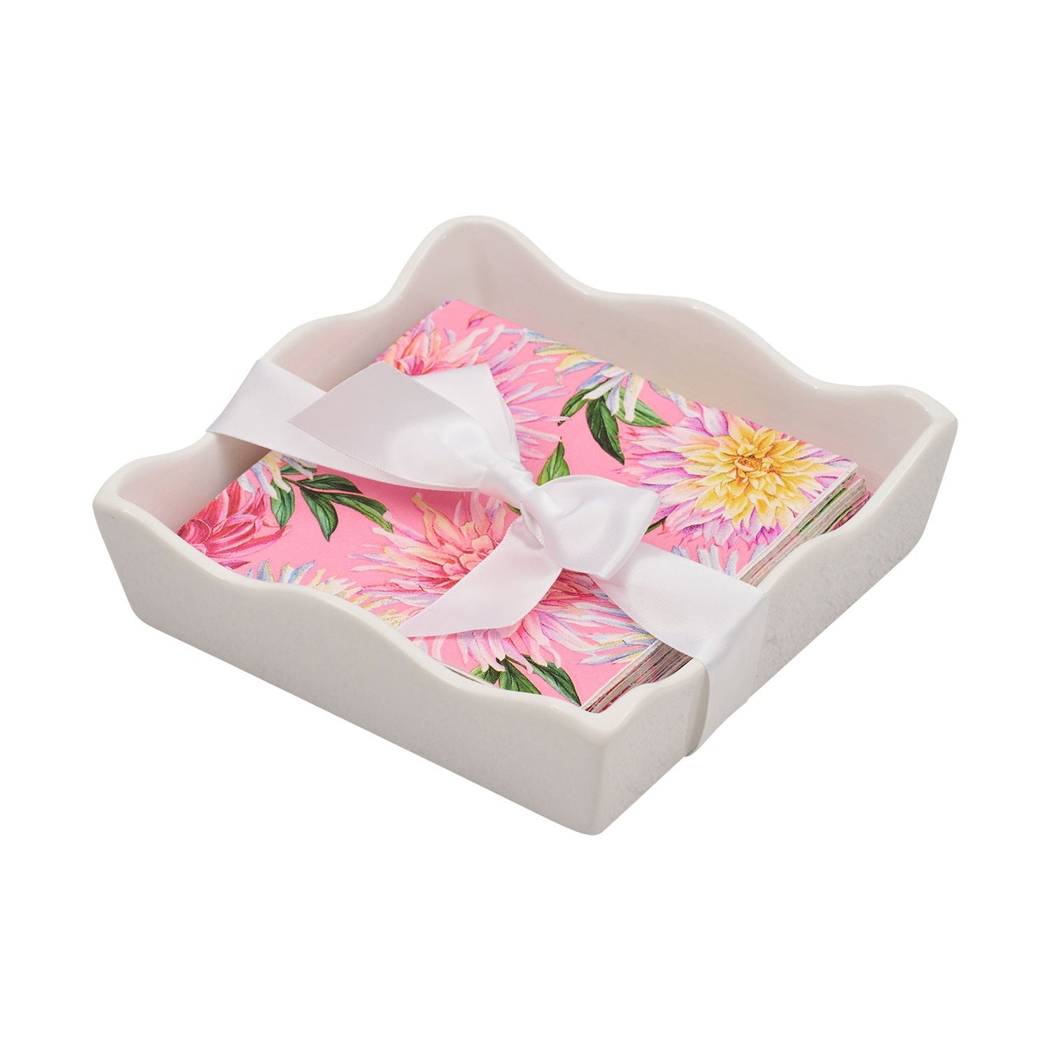 White ceramic napkin holder with a wavy edge filled with pink floral paper napkins tied with a white satin ribbon featuring vibrant pink and yellow dahlia patterns