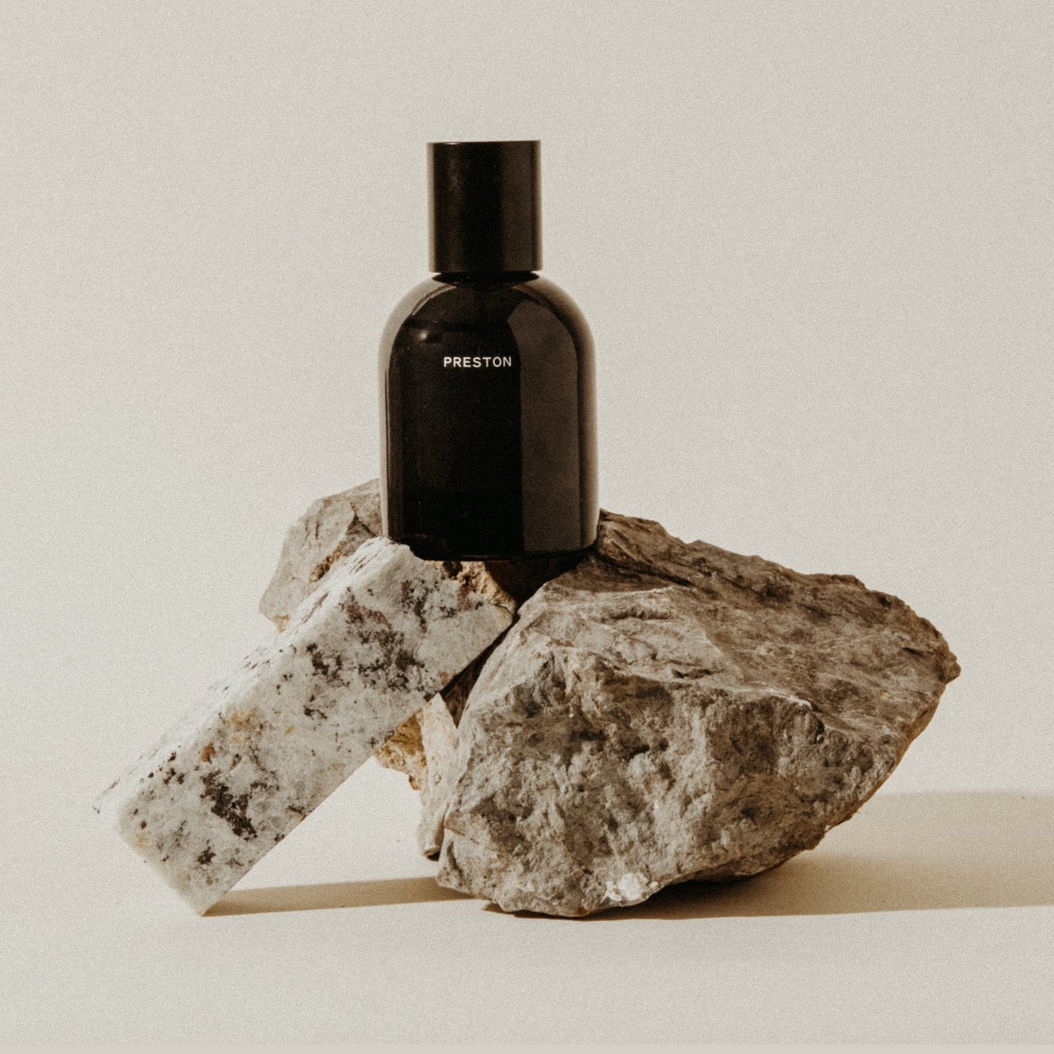 A black Preston fragrance bottle sits atop a stack of natural rocks including a speckled white granite piece and a dark gray textured stone on a light beige background