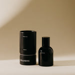 Still life photograph featuring a sleek black Preston Amber Cologne bottle alongside its cylindrical packaging with white text against a minimalist beige backdrop