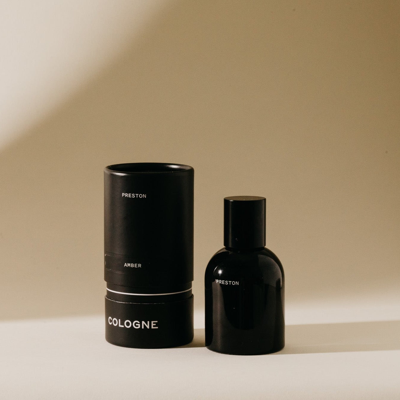 Still life photograph featuring a sleek black Preston Amber Cologne bottle alongside its cylindrical packaging with white text against a minimalist beige backdrop