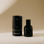 A sleek black Preston Leather Cologne bottle stands next to its matching cylindrical packaging featuring white text, all set on a warm beige background with subtle shadows