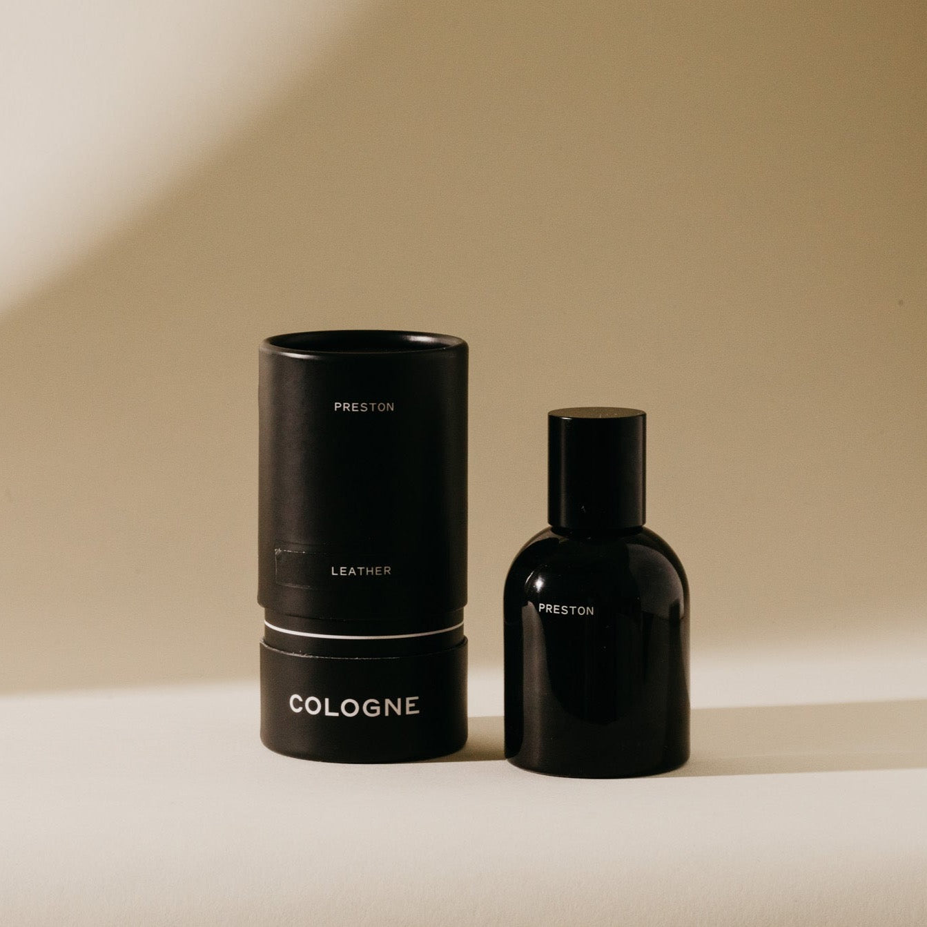 A sleek black Preston Leather Cologne bottle stands next to its matching cylindrical packaging featuring white text, all set on a warm beige background with subtle shadows