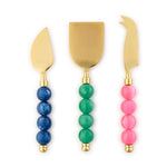 Set of three gold-toned cheese knives featuring a curved spreader, wide spade, and serrated pronged knife, each with distinct beaded handles in vibrant blue, green, and pink
