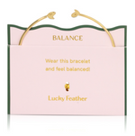 Gold open cuff bracelet with stylized bird ends on a pink "Lucky Feather" brand card featuring a green wavy border and "Balance" messaging