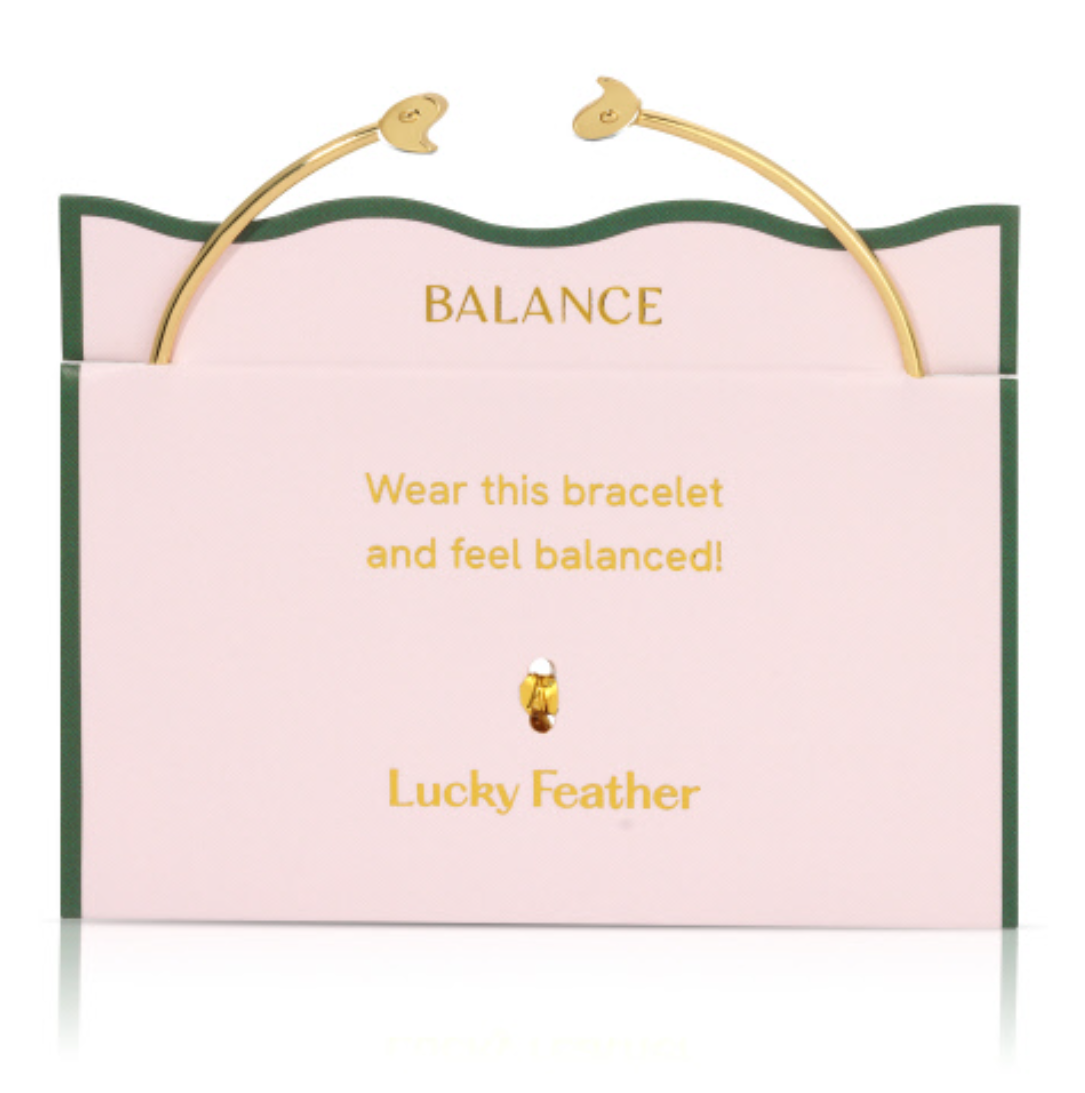 Gold open cuff bracelet with stylized bird ends on a pink "Lucky Feather" brand card featuring a green wavy border and "Balance" messaging