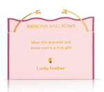 Gold Lucky Feather bracelet with two delicate bow charms presented on a light pink card with a wavy dark pink border, displaying gold text 'RIBBONS AND BOWS' and 'Wear this bracelet and know you're a true gift' above a tiny feather emblem