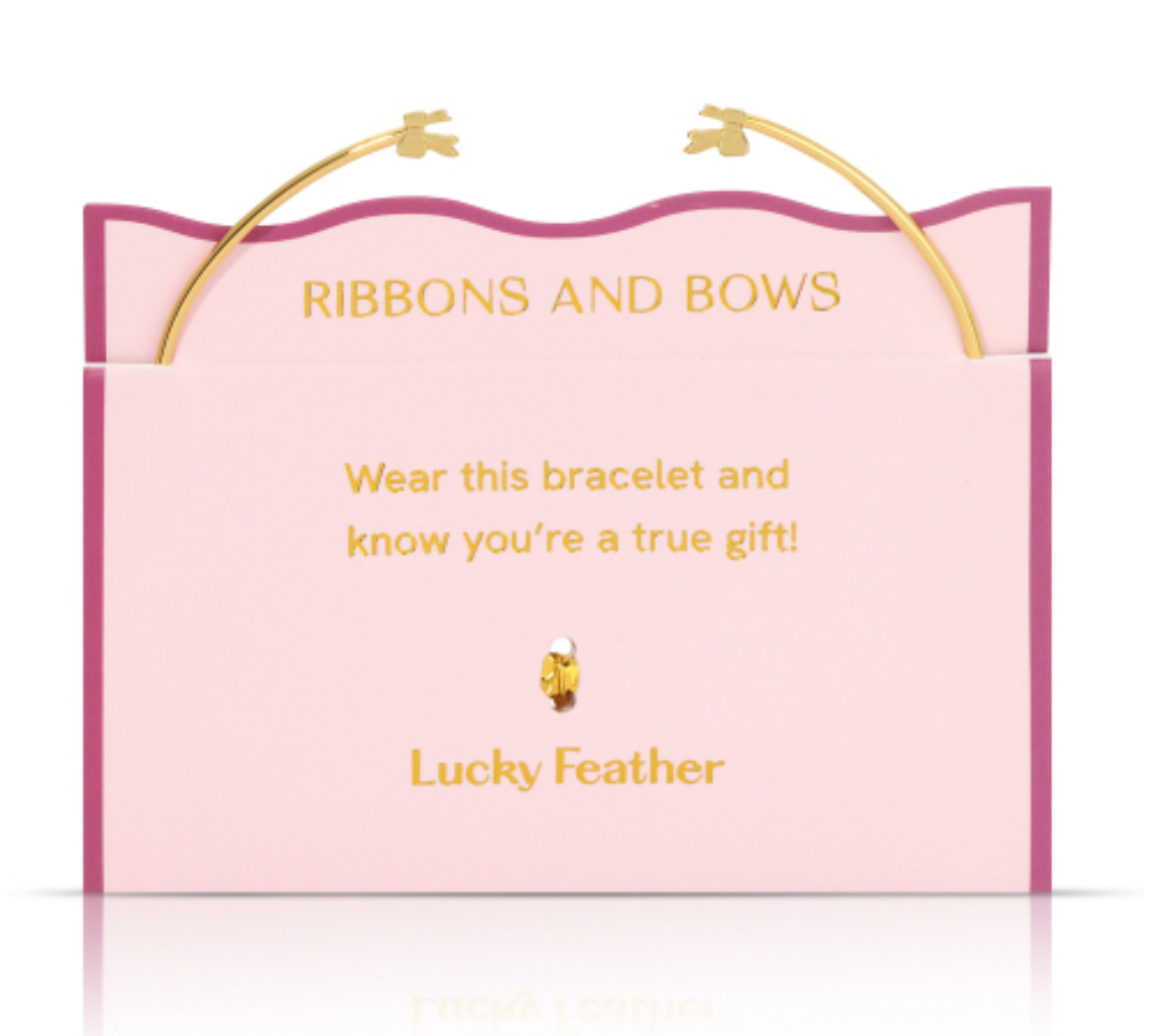 Gold Lucky Feather bracelet with two delicate bow charms presented on a light pink card with a wavy dark pink border, displaying gold text 'RIBBONS AND BOWS' and 'Wear this bracelet and know you're a true gift' above a tiny feather emblem