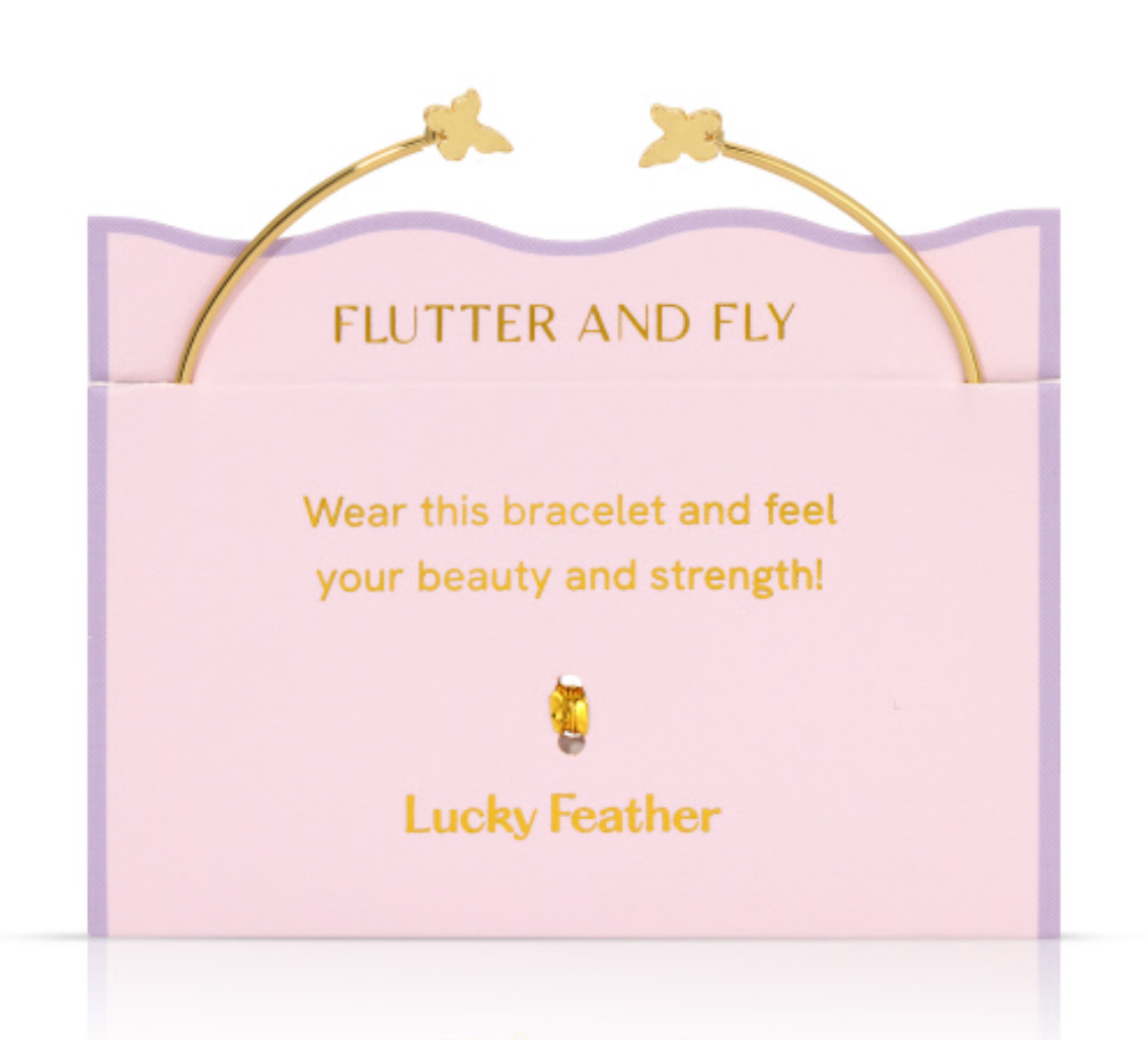 Product photography Lucky Feather gold butterfly bangle bracelet resting on a pink wavy-edged display card featuring Flutter and Fly text and a small golden bead charm