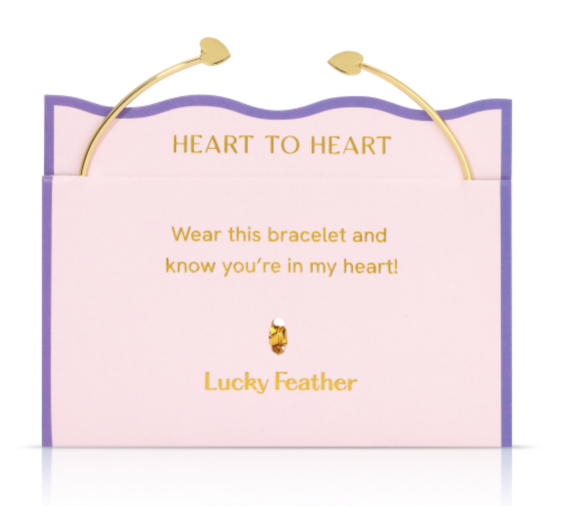 Lucky Feather product photo of a gold-tone Heart to Heart open cuff bracelet with small heart ends displayed on a pink and lavender card featuring the golden text HEART TO HEART and the message Wear this bracelet and know you're in my heart