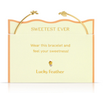A gold open-cuff bracelet with delicate bee and feather charms presented on a pale yellow product card with wavy orange trim, gold lettering including Sweetest Ever and Lucky Feather, and a small sparkling amber-colored gem