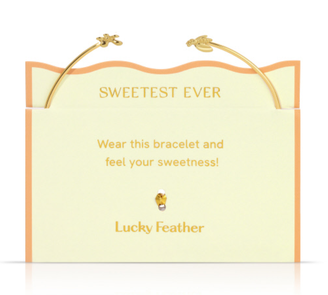 A gold open-cuff bracelet with delicate bee and feather charms presented on a pale yellow product card with wavy orange trim, gold lettering including Sweetest Ever and Lucky Feather, and a small sparkling amber-colored gem