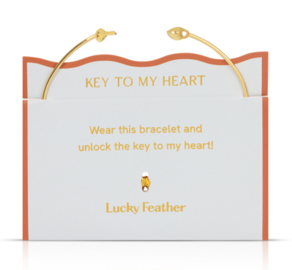 Product image by Lucky Feather of a gold cuff bracelet with key ends presented on a white and orange "KEY TO MY HEART" display card