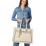 A smiling woman in a denim shirt and jeans on a white background holds a natural canvas tote bag featuring a black line drawing of a Birkin handbag outline