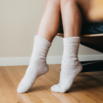 Photograph of legs and feet in fuzzy cream socks on a light wood floor, one foot elevated, with a dark chair and beige wall background