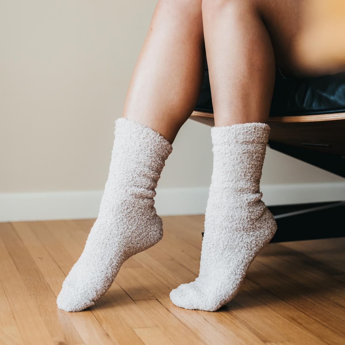 Photograph of legs and feet in fuzzy cream socks on a light wood floor, one foot elevated, with a dark chair and beige wall background