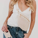 A woman in a white tank top and light wash jeans wearing a beige woven fanny pack with gold zippers and holding a matching coin purse, accessorized with a gold cross necklace