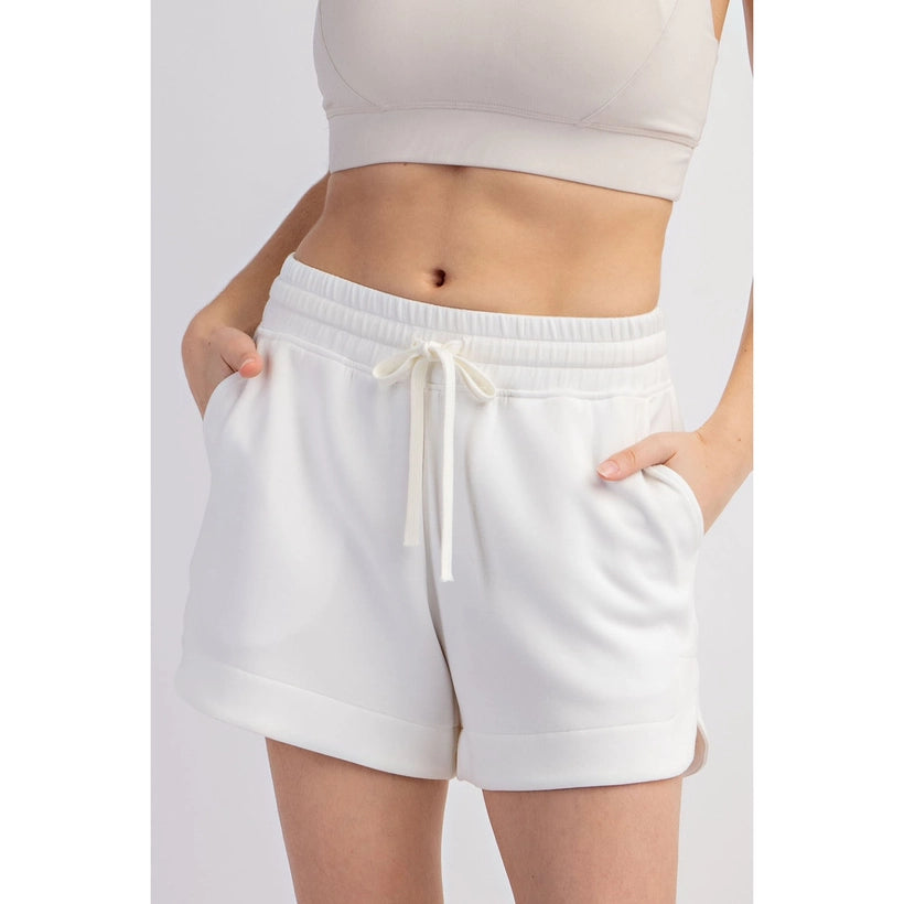 A person wearing a cream V-neck sports bra and matching fleece shorts with a drawstring waist and rolled cuffs, hands in pockets