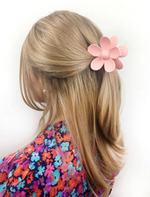 A close-up of a person with blonde hair styled in a half-updo, adorned with a large matte pink flower claw clip, wearing a vibrant floral print top featuring blue, orange, and purple flowers