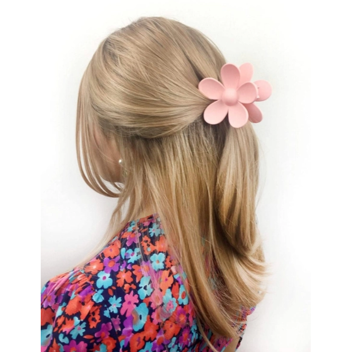 A close-up of a person with blonde hair styled in a half-updo, adorned with a large matte pink flower claw clip, wearing a vibrant floral print top featuring blue, orange, and purple flowers