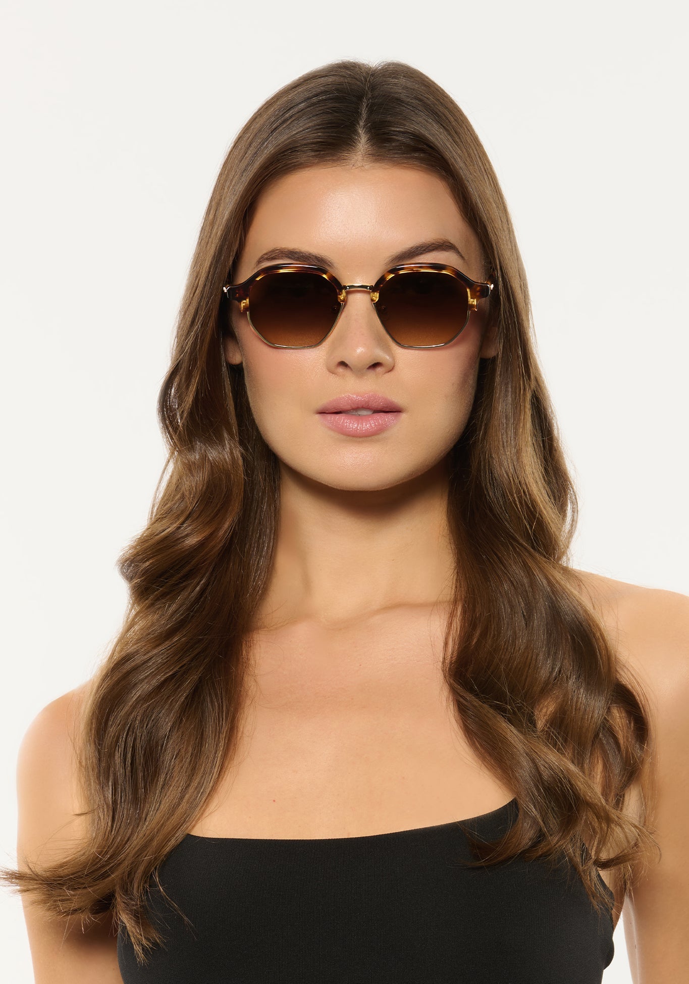 Studio photograph of a woman with long wavy brunette hair wearing stylish brown tortoise-shell hexagonal sunglasses with gradient lenses and a black strapless top against a white background