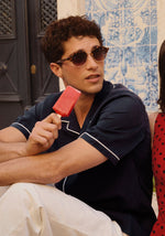 Photograph of a young man wearing tortoiseshell sunglasses and a navy blue shirt with white piping holding a vibrant red popsicle against a backdrop of intricate blue and white Portuguese azulejo tiles and a dark carved door