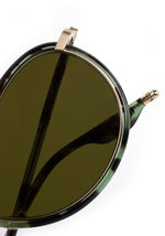 Close-up of round dark green lens sunglasses with a green tortoiseshell frame, featuring an ornate gold volute hinge and "DESIGNED IN NEW ORLEANS" inscribed on the inner temple
