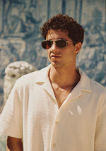 A young man with dark curly hair and tortoise-shell sunglasses wears a cream textured collared shirt and silver chain against a backdrop of blurry blue tiled patterns and a white classical statue