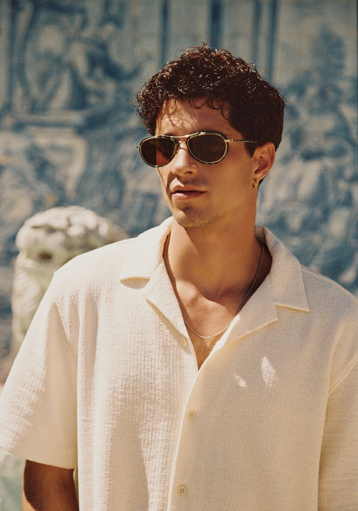 A young man with dark curly hair and tortoise-shell sunglasses wears a cream textured collared shirt and silver chain against a backdrop of blurry blue tiled patterns and a white classical statue