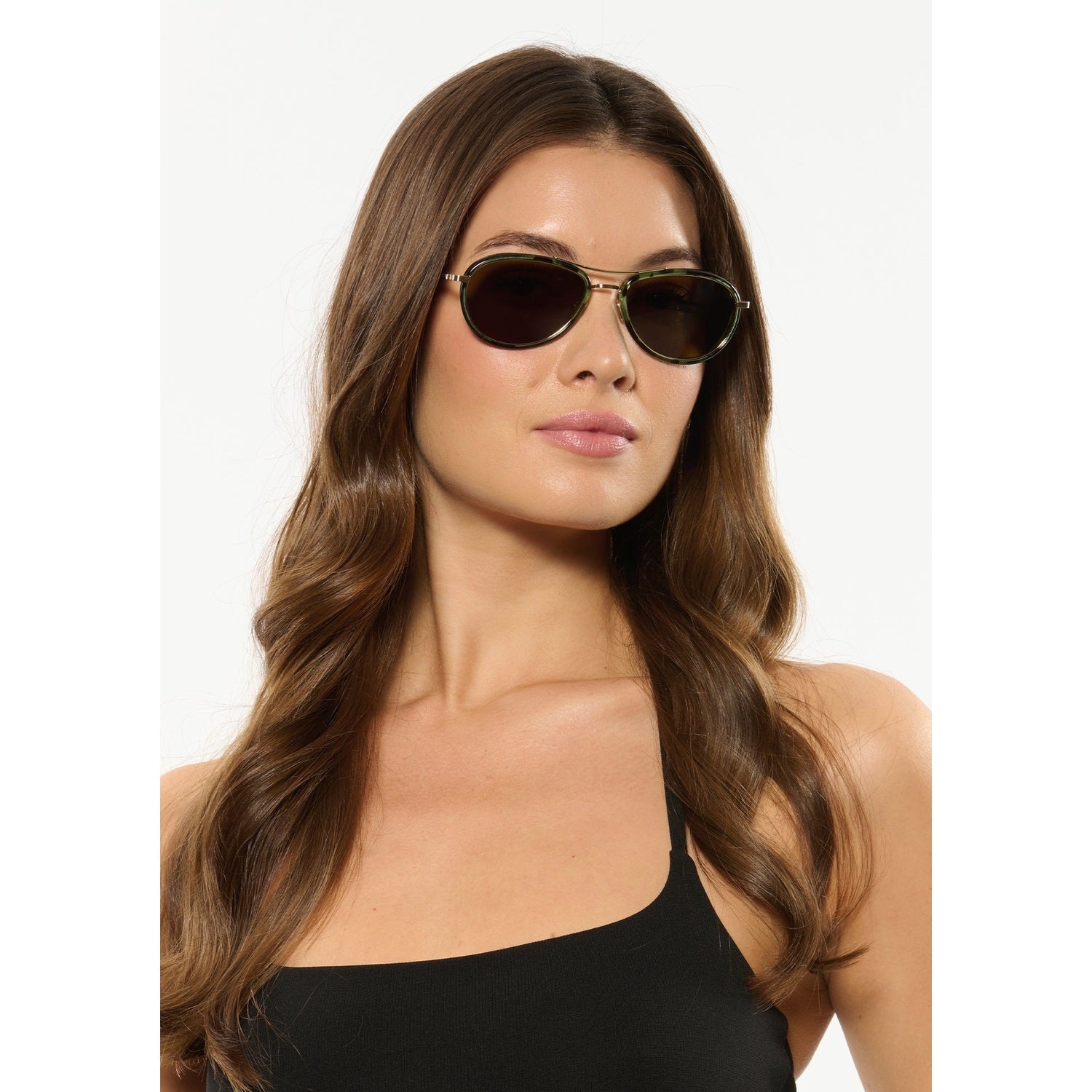 Photography of a woman posing in a studio with long wavy brown hair, wearing green tortoiseshell aviator sunglasses, glossy pink lips, and a black tank top