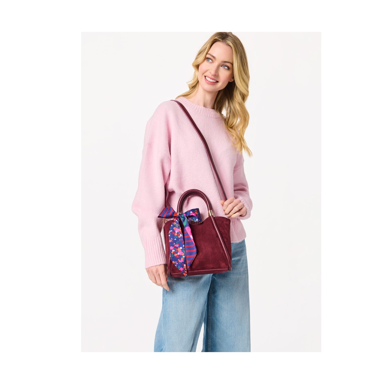 Woman wearing a pink sweater and blue jeans holding a burgundy handbag with a floral scarf tied to the handle