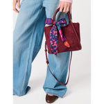 Person holding a burgundy suede handbag with a floral and striped scarf tied to the handle