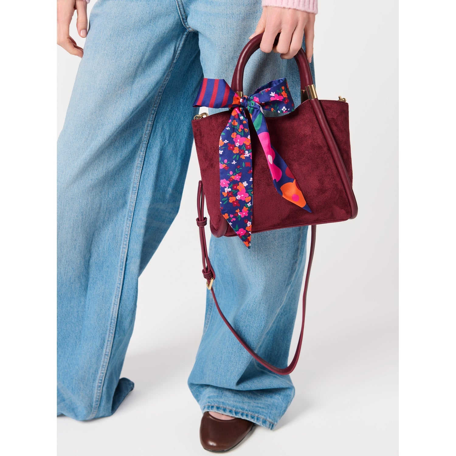 Person holding a burgundy suede handbag with a floral and striped scarf tied to the handle