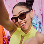 Photograph of a smiling young woman taking a selfie wearing black goodr sunglasses and a bright yellow top against a vibrant background of pink orange and blue shiny streamers