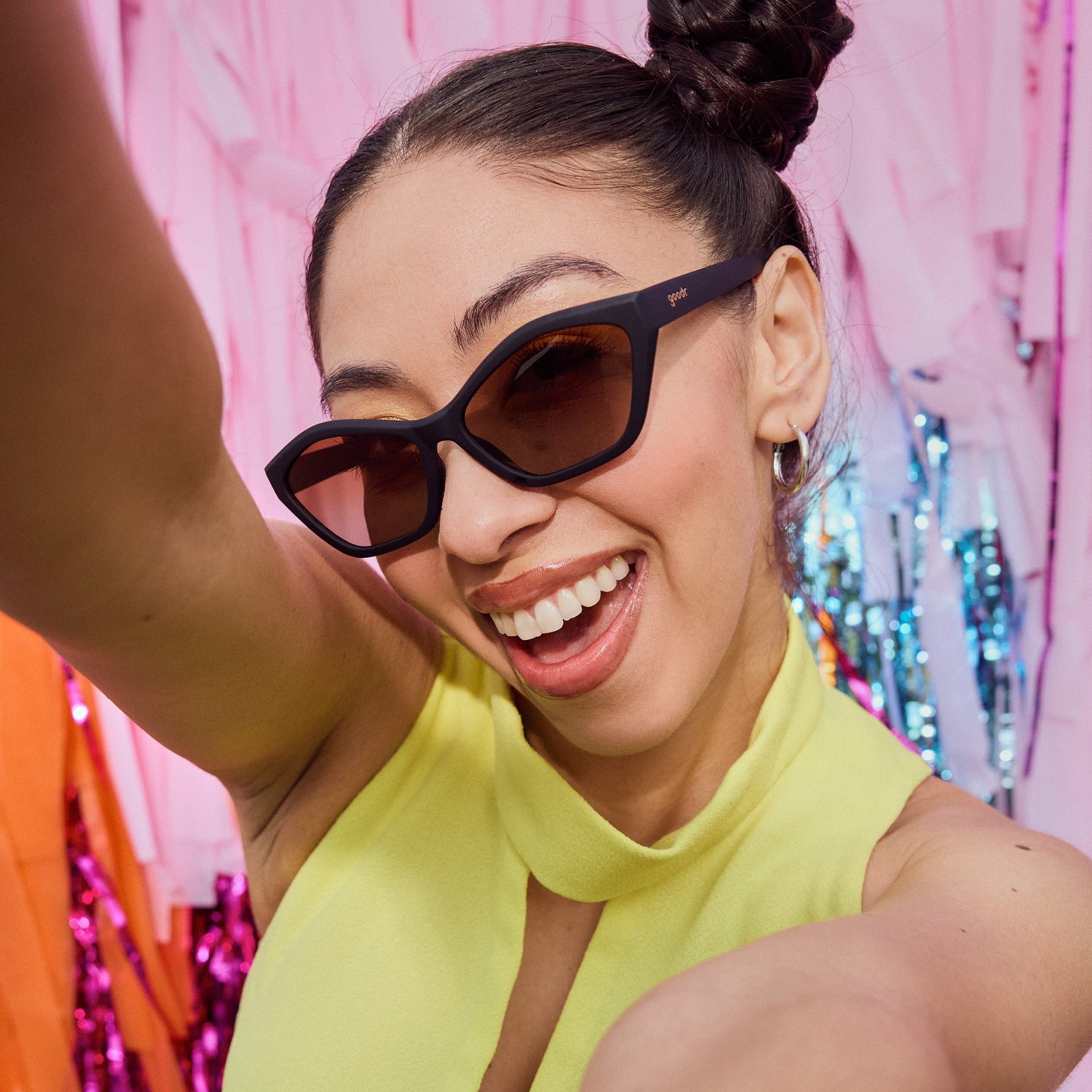 Photograph of a smiling young woman taking a selfie wearing black goodr sunglasses and a bright yellow top against a vibrant background of pink orange and blue shiny streamers