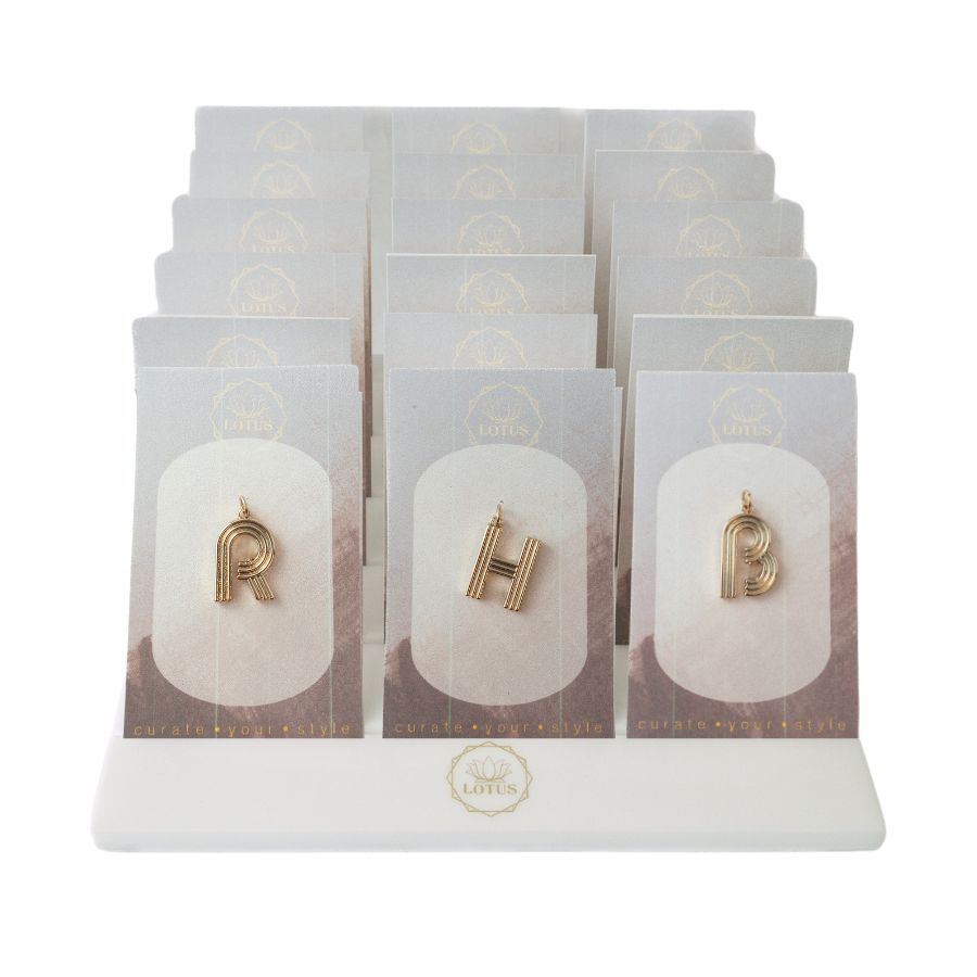 Product photography by Lotus of gold initial pendants on a white tiered display stand featuring ribbed letter charms for R H and B on neutral cards with a gold lotus logo and curate your style branding