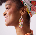 Photograph of a smiling woman looking left, wearing a colorful fruit-patterned headwrap, vibrant beaded fringe earrings with pineapple and watermelon designs, and a coral beaded necklace
