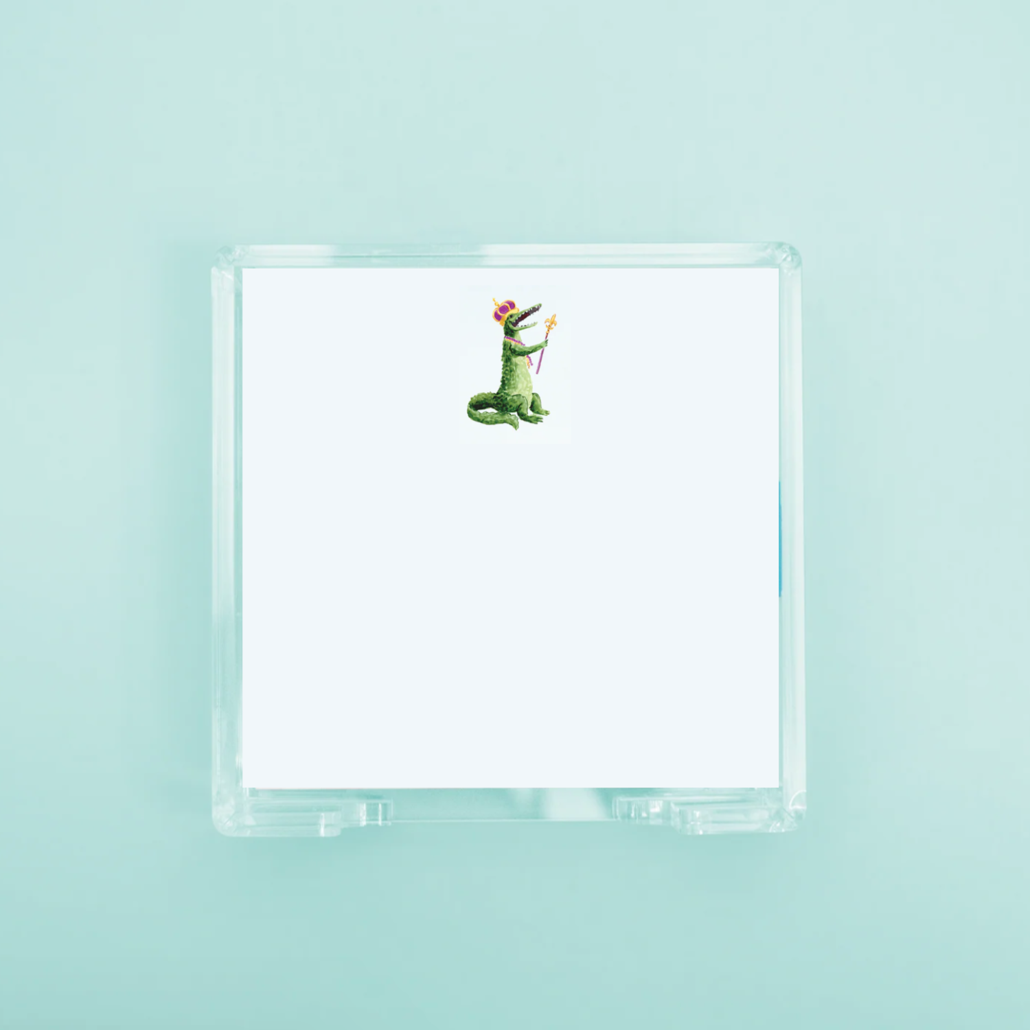 A stack of white note paper in a clear acrylic holder featuring a cartoon green alligator illustration wearing a purple and gold crown and holding a scepter against a light blue background