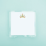 Illustration of a gold crown with vibrant purple and green jewels on a white notepad paper in a clear acrylic holder against a soft blue-green backdrop