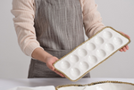 Person in a beige ribbed shirt and striped gray apron holding a white ceramic deviled egg platter with a decorative gold beaded rim and twelve indentations